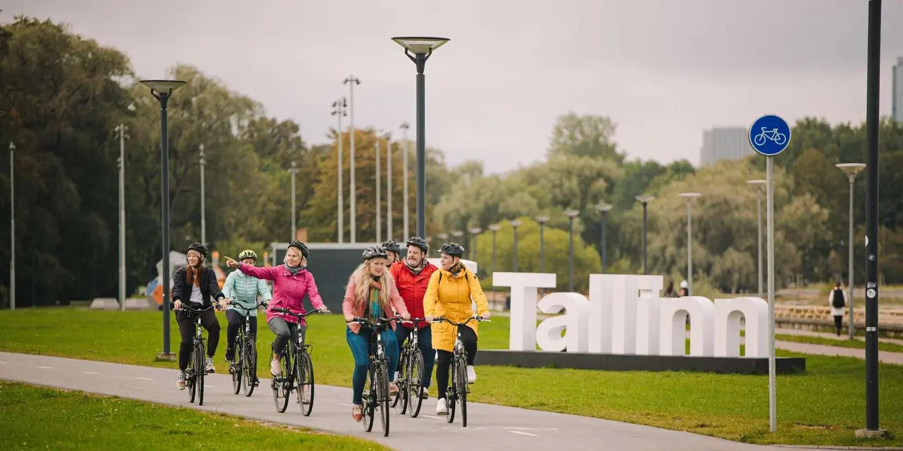 Seven cyclists in helmets and bright rain jackets riding along a park path past a large white Tallinn sign, one pointing ahead