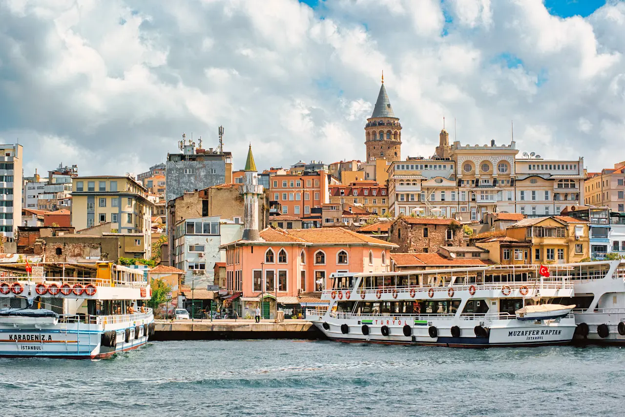 Karaköy's waterfront on the Golden Horn, with white passenger ferries moored in front of colourful buildings and Galata Tower rising above them under a bright, cloudy sky