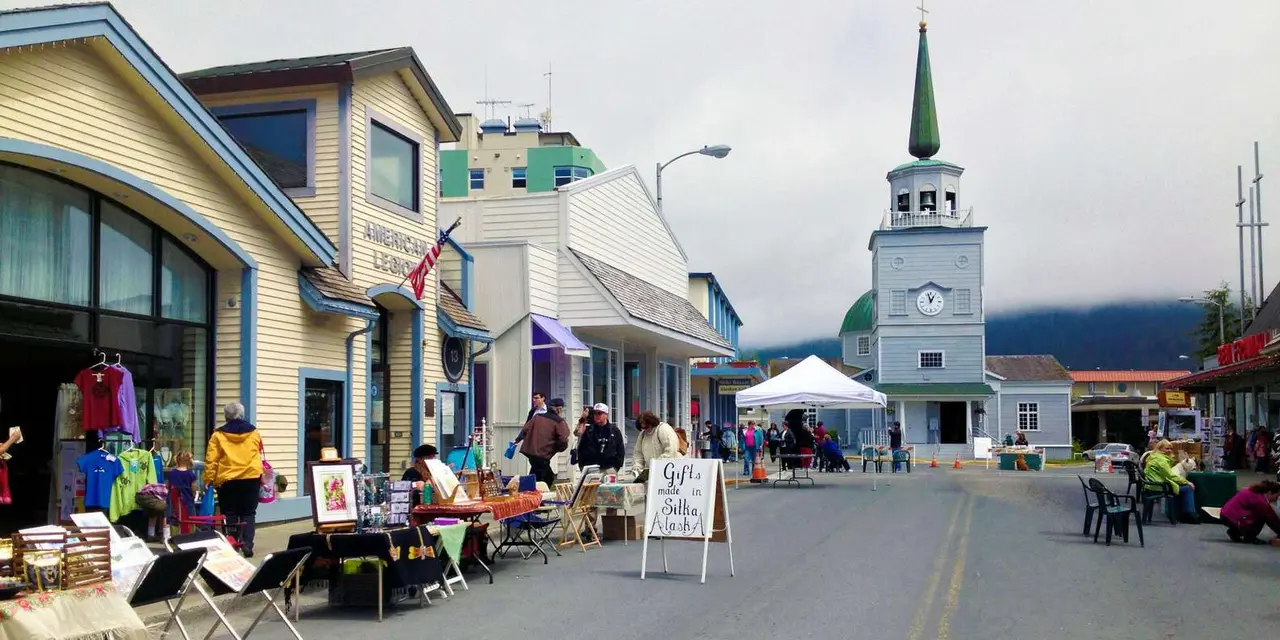 A downtown Sitka street under low cloud, with craft stalls on the pavement and a church tower with a green spire and clock at the end of the block