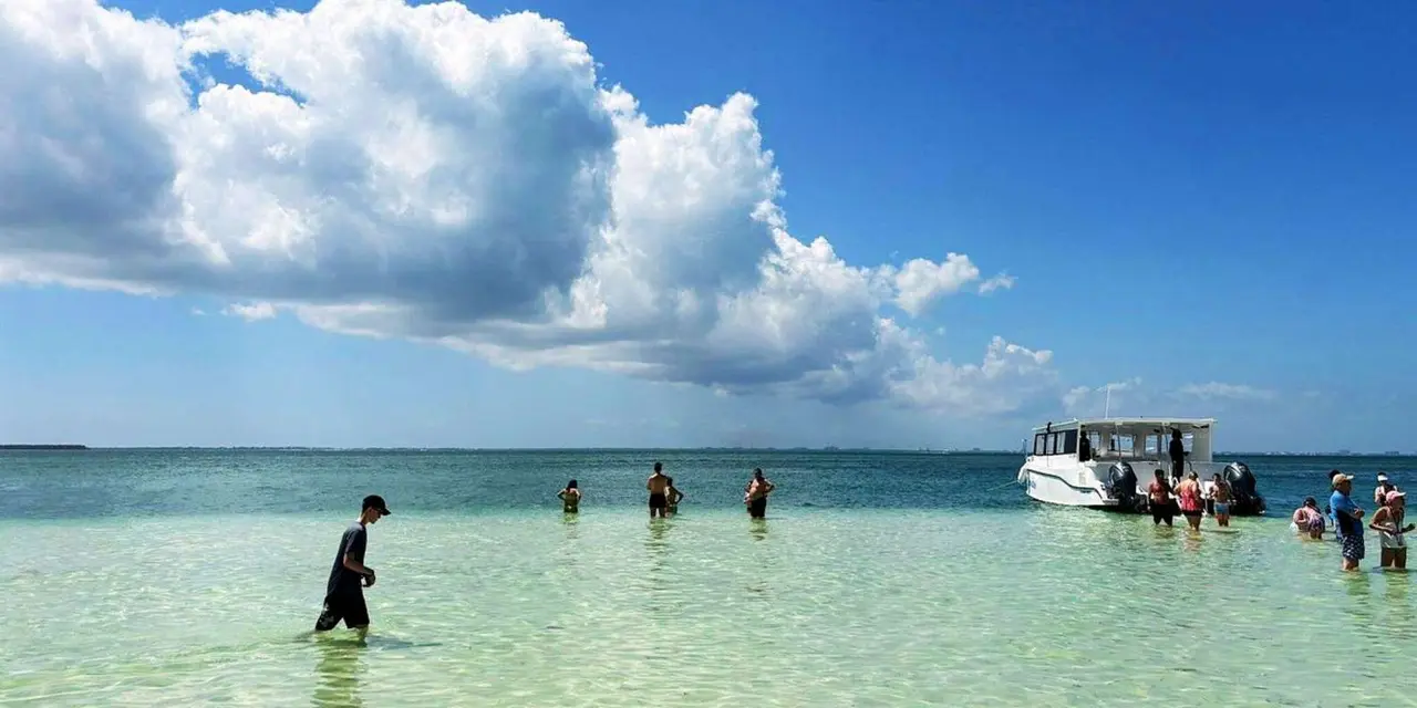 People wading in knee-deep clear water off a sandy shore under tall white clouds, with a small tour boat to the right