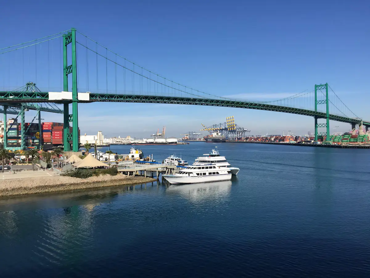 The green Vincent Thomas Bridge spanning the Los Angeles Harbor main channel in San Pedro under a clear blue sky, a white harbor-cruise boat moored below
