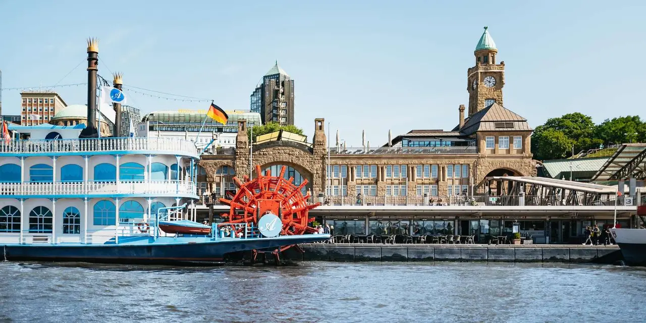 The brick-and-sandstone St. Pauli Landungsbrücken harbor pier building with its clock tower and a white paddle steamer with red paddle wheel beside it