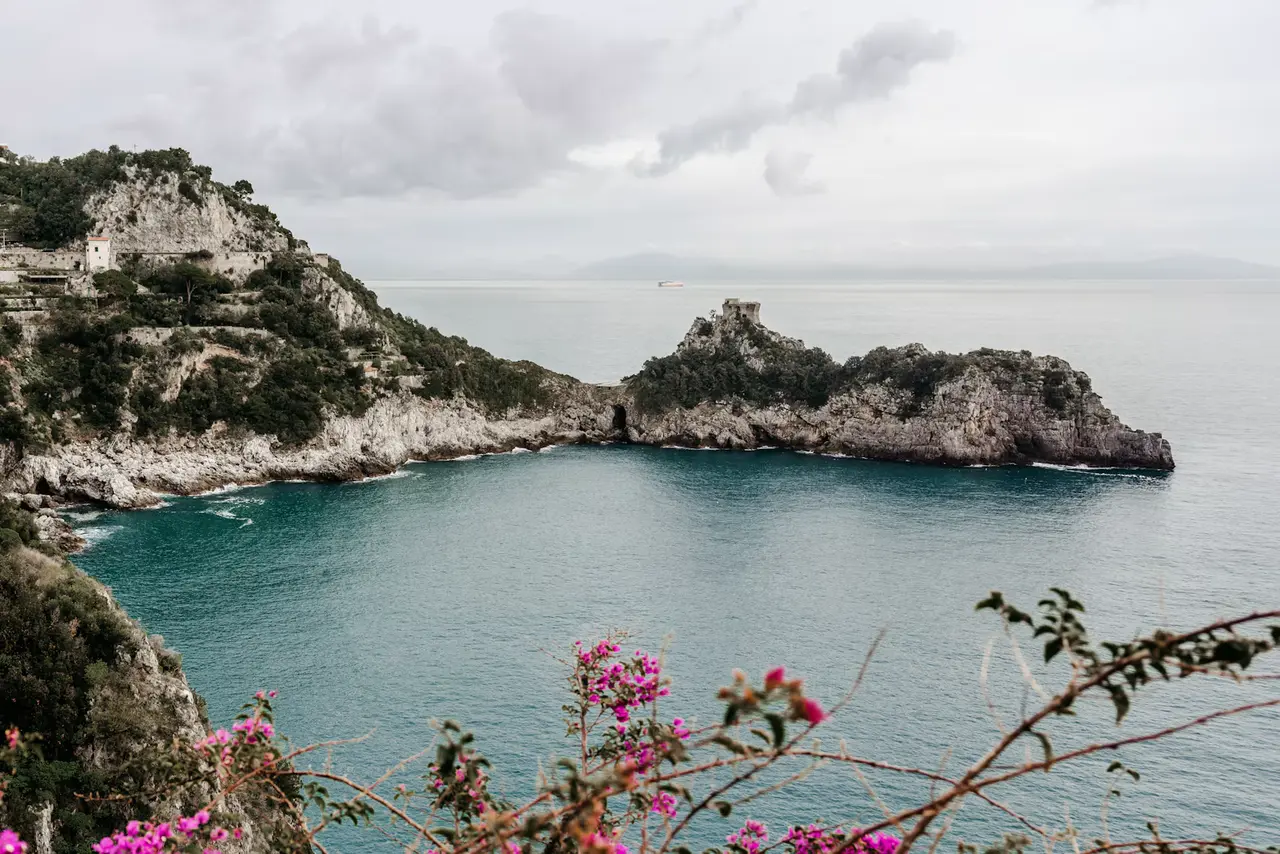 A turquoise cove between rocky, green headlands on the Amalfi Coast, which begins just west of Salerno, with pink bougainvillea in the foreground