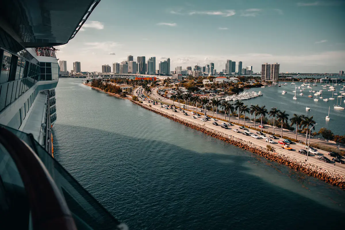 View from a cruise ship's deck down the PortMiami channel: the palm-lined MacArthur Causeway, a marina full of boats and the downtown Miami skyline under a blue sky