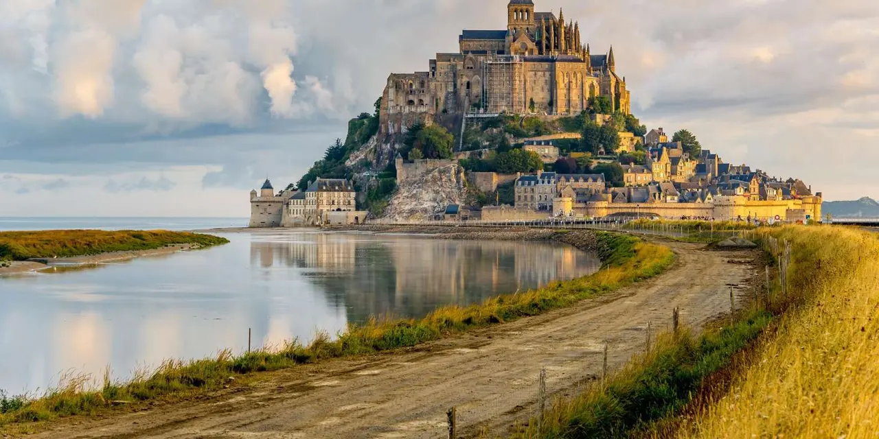 Mont Saint-Michel's abbey and village on its rock in warm evening light, with a tidal channel and a dirt track in the foreground