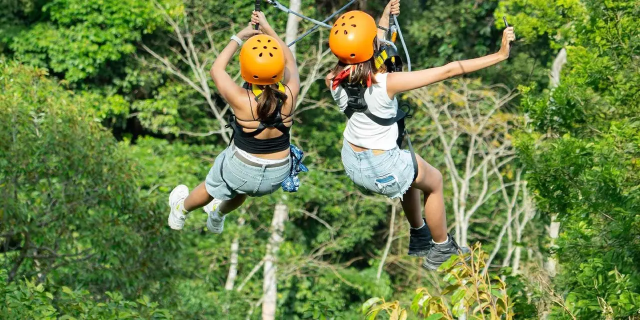 Two riders in orange helmets on a zipline among jungle trees