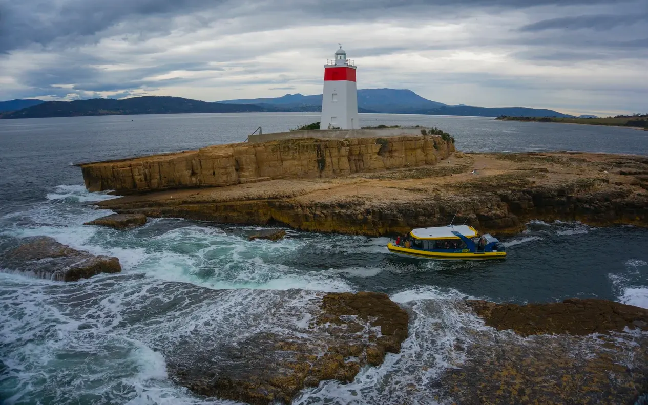 A red-and-white lighthouse on a sandstone islet with a yellow boat in the swell below
