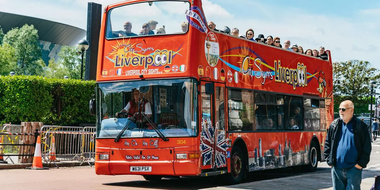 A red open-top City Sights bus with passengers on the upper deck, parked beside a green hedge near the Royal Albert Dock on a sunny day