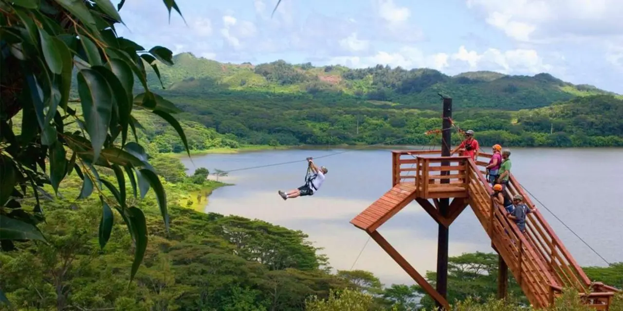 A rider in a harness flying along a zipline away from a tall wooden platform above a green-rimmed reservoir