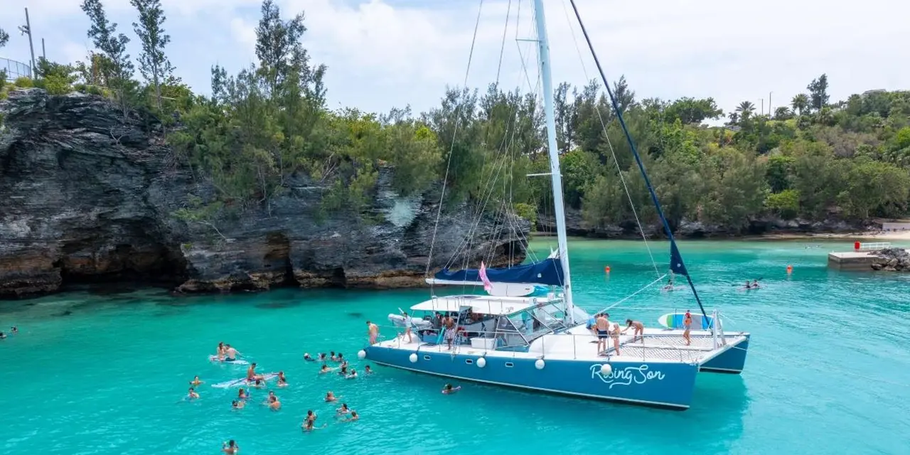 A sailing catamaran anchored in bright turquoise water off a green shoreline, with swimmers around it