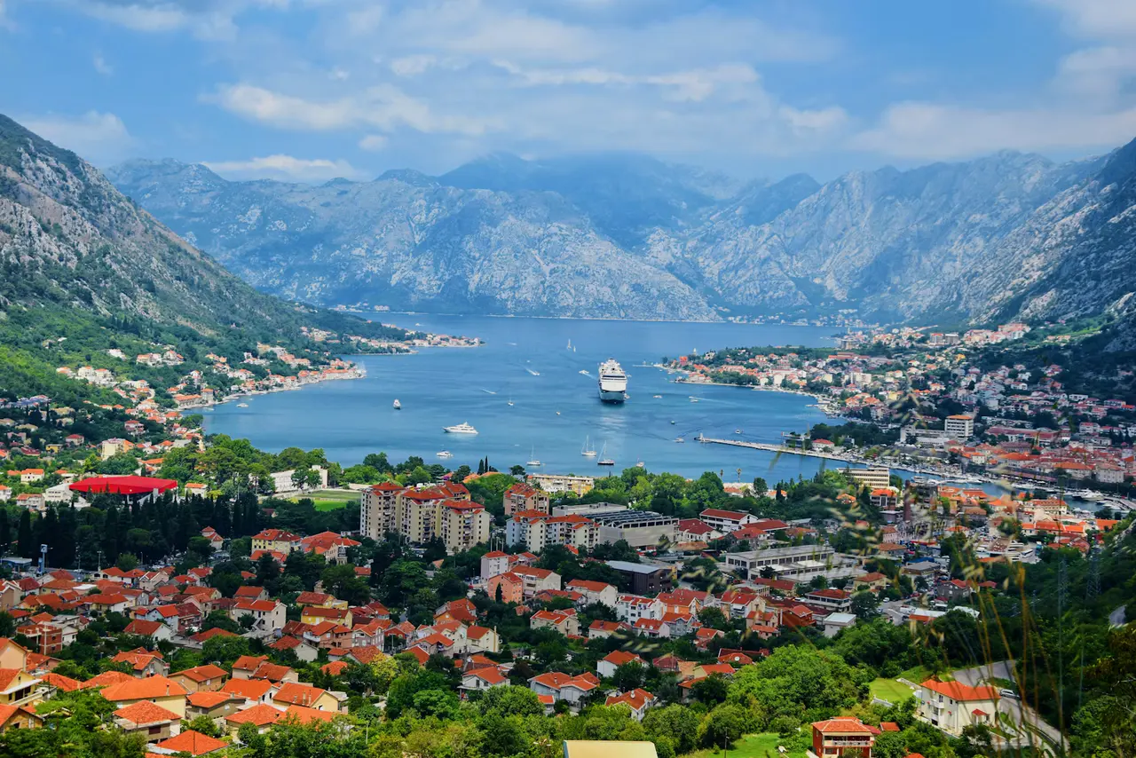 A white cruise ship at anchor in the blue Bay of Kotor, seen from the hillside above the terracotta rooftops of the old town, with steep mountains rising behind it