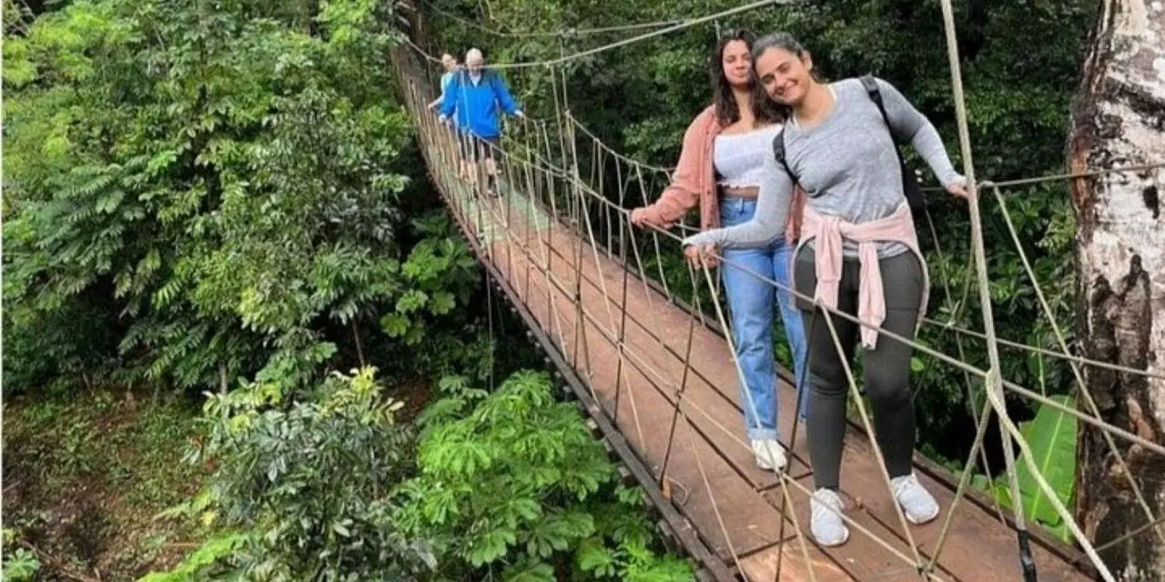 Two women in light jackets tied at the waist smile on a rope suspension bridge in dense green jungle, with other visitors crossing behind them
