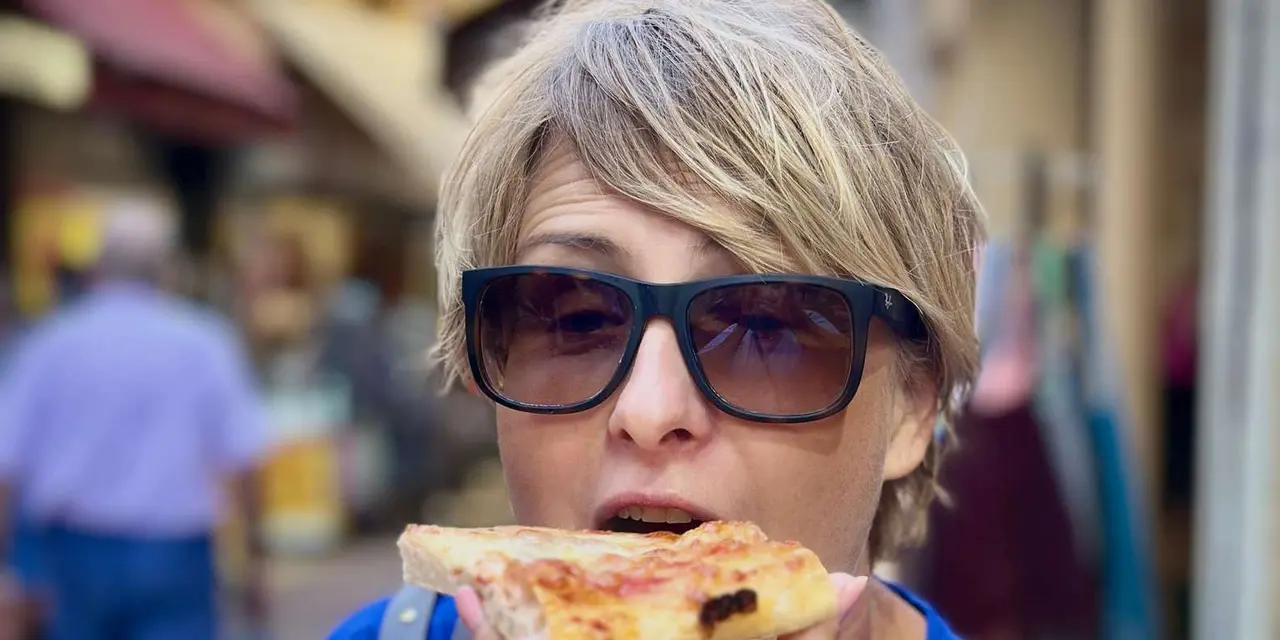 A woman in sunglasses biting into a slice of street food in a narrow old-town lane