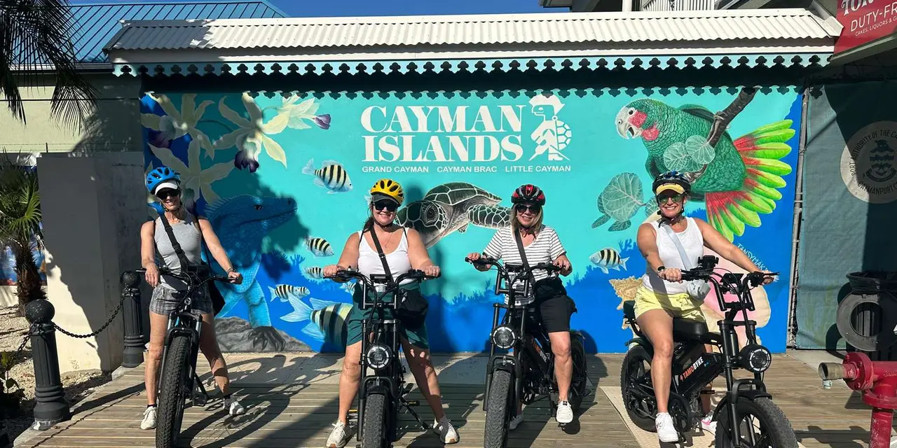 Four riders in helmets on fat-tire e-bikes in front of a blue Cayman Islands mural painted with fish and parrots