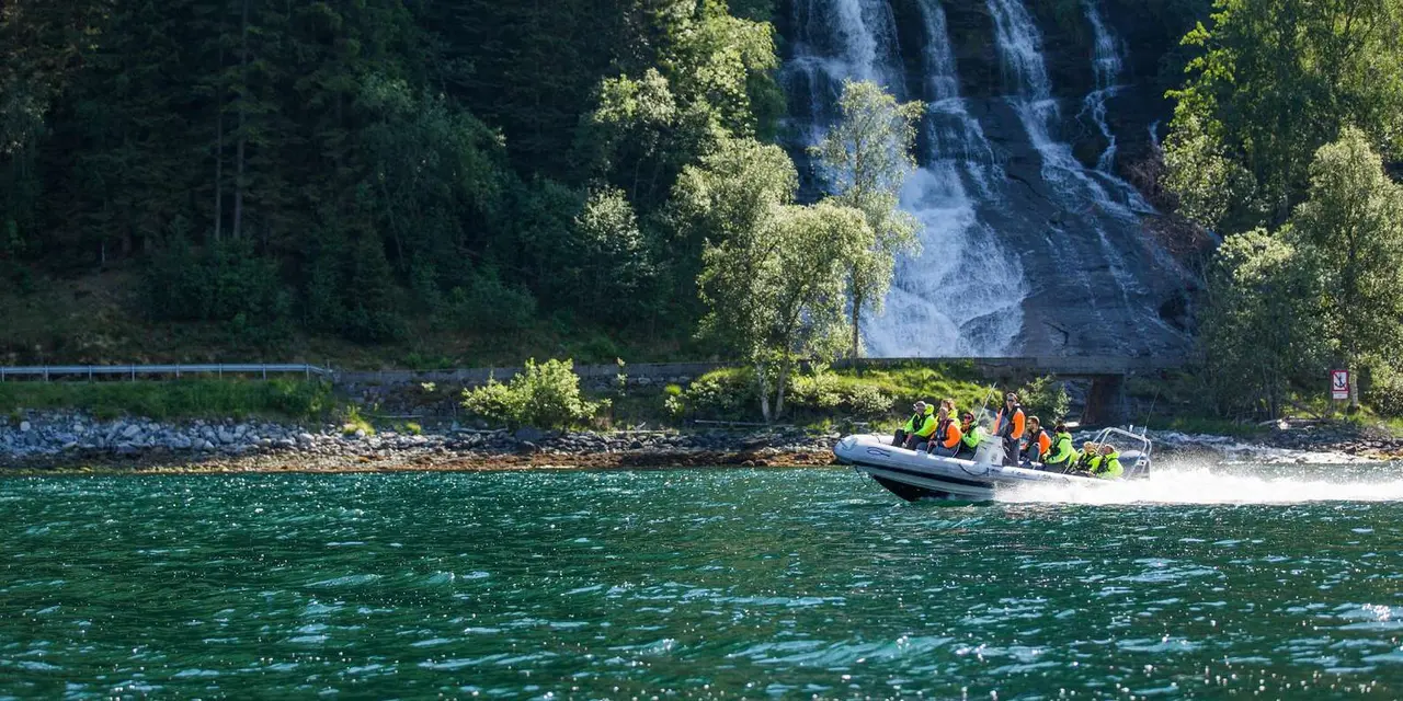 A RIB boat full of passengers in bright suits on green fjord water below a tall waterfall