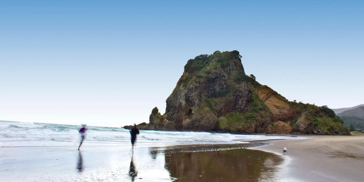 Lion Rock rising from Piha beach, with a walker on wet black sand reflecting the sky