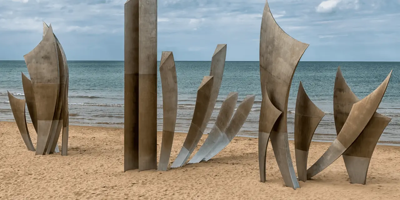 Tall brushed-steel memorial sculptures standing on a sandy beach with the sea behind