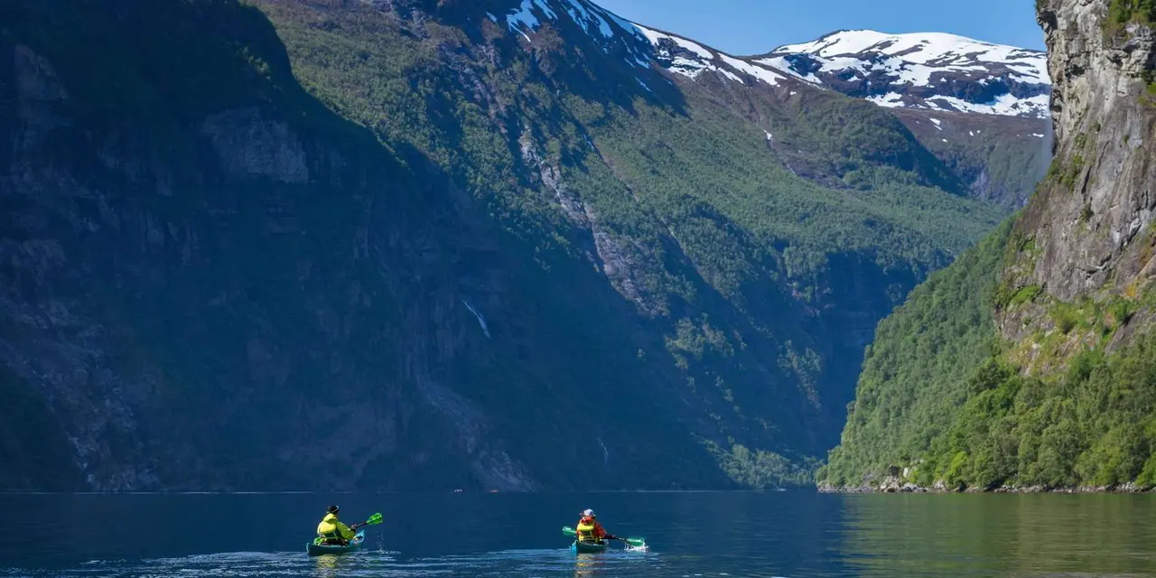 A lone kayaker on dark fjord water between steep green mountainsides and snowy peaks