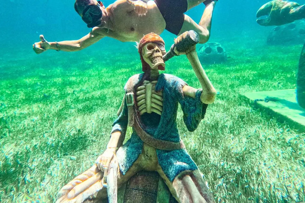 A snorkeler swimming above an underwater statue of a skeleton pirate on a bed of seagrass