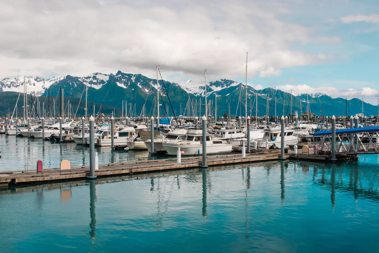 Fishing boats and sailboats moored in the turquoise water of Seward's Small Boat Harbor, with the snow-capped Kenai Mountains behind