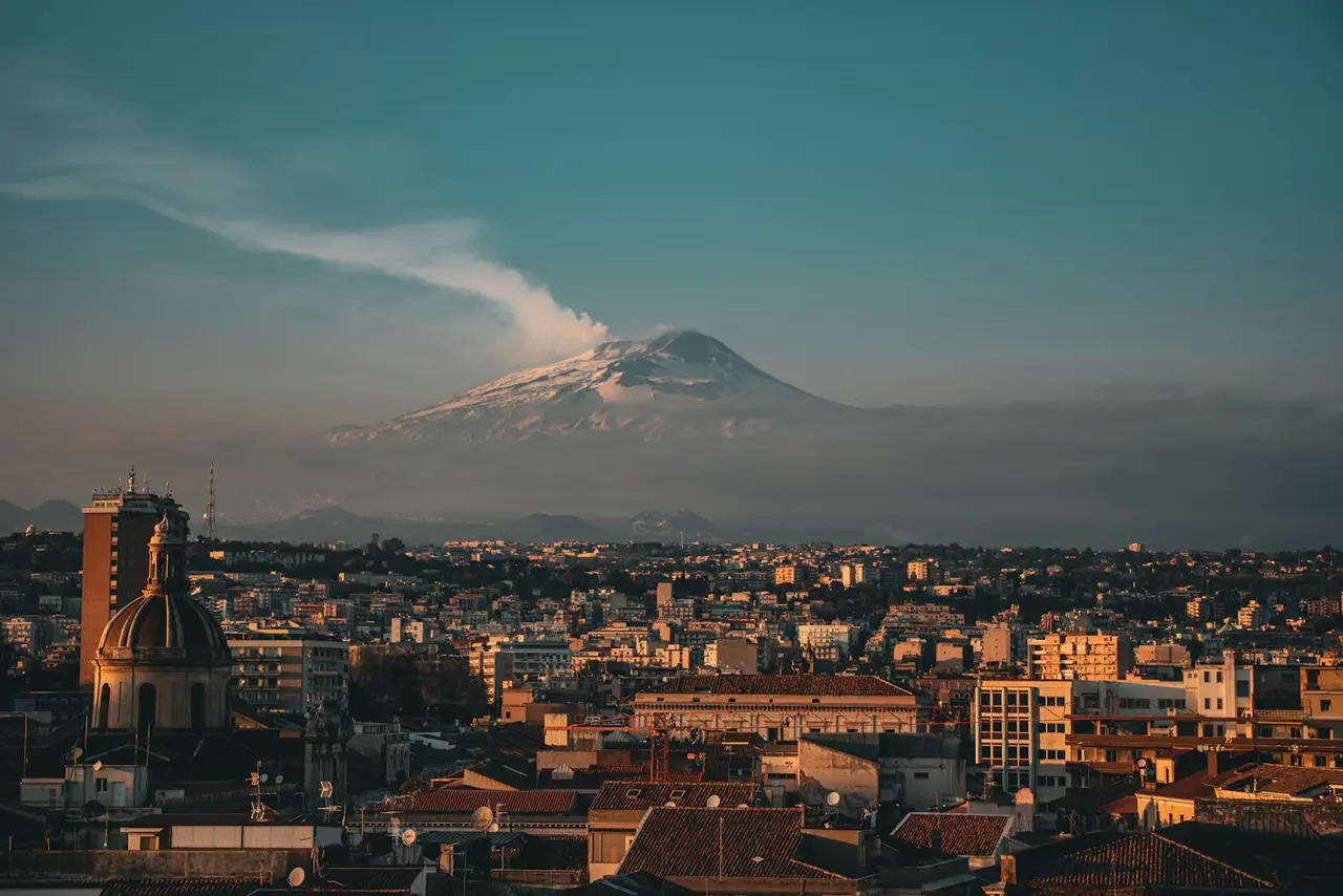 Mount Etna's snow-dusted cone with a plume of steam rising behind the rooftops and domes of Catania at golden hour, with clear sky in the upper left