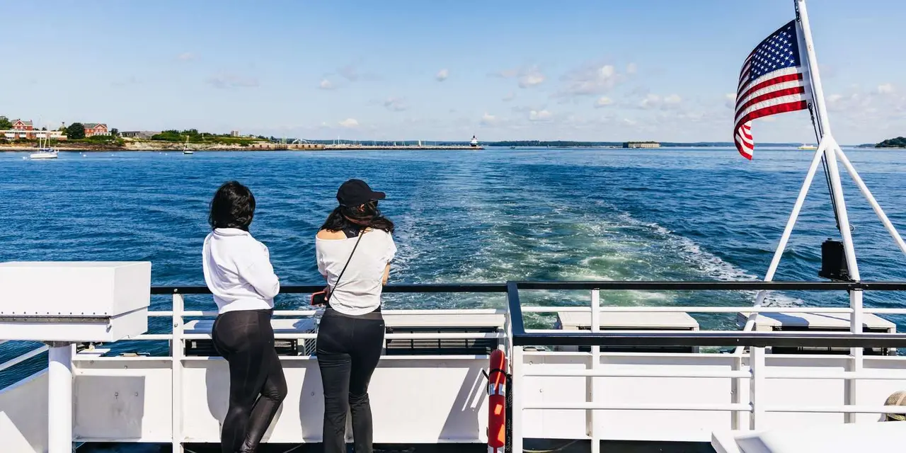 Two passengers at the stern rail of a cruise boat looking back at its wake and the distant Casco Bay shoreline