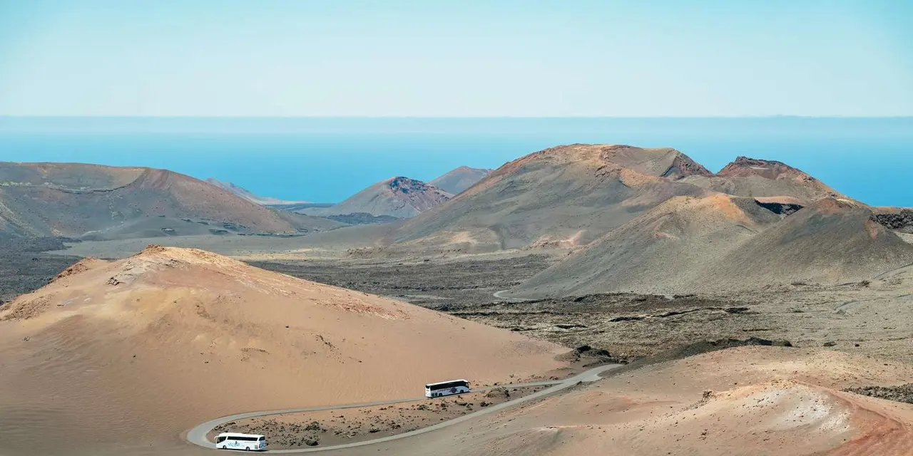 A road winding between ochre and red volcanic cones, with a coach on it and the sea beyond