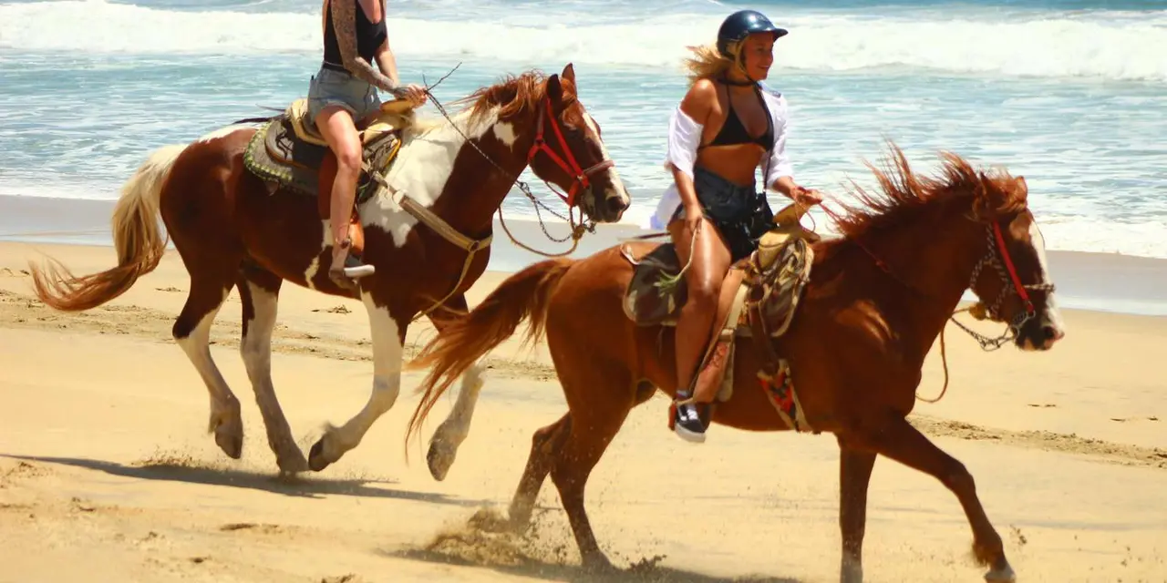 Two riders in helmets on chestnut horses cantering along a sandy beach with waves behind them