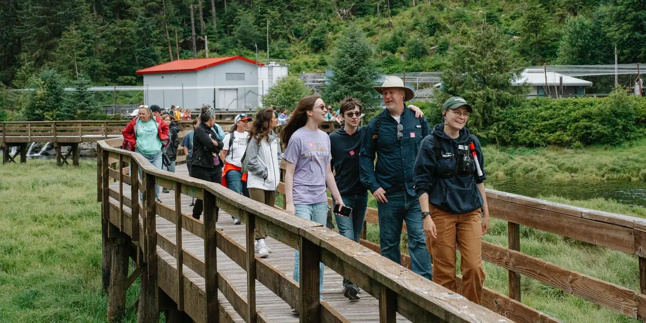 A tour group on a wooden boardwalk through forest, a red-roofed building behind