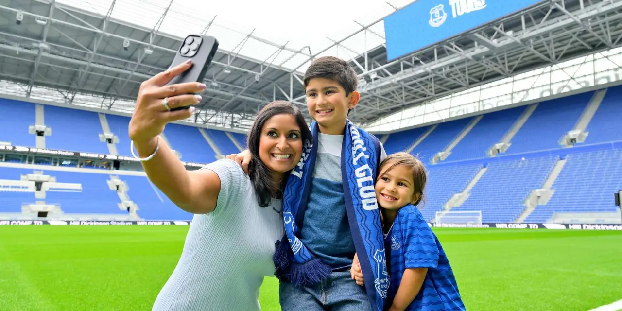 A mother taking a selfie with two children in Everton scarves and shirts on the pitch of Hill Dickinson Stadium, rows of blue seats behind them