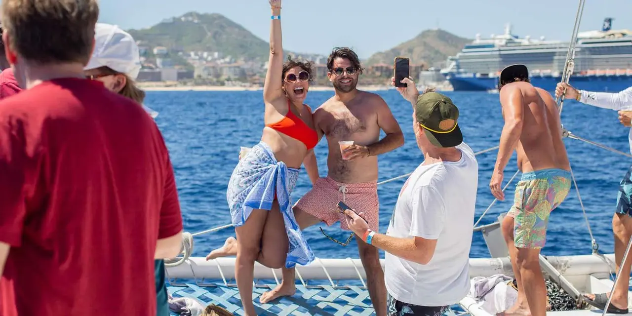 Passengers on the deck of a catamaran in Cabo's bay, one woman posing at the rail with a cruise ship behind