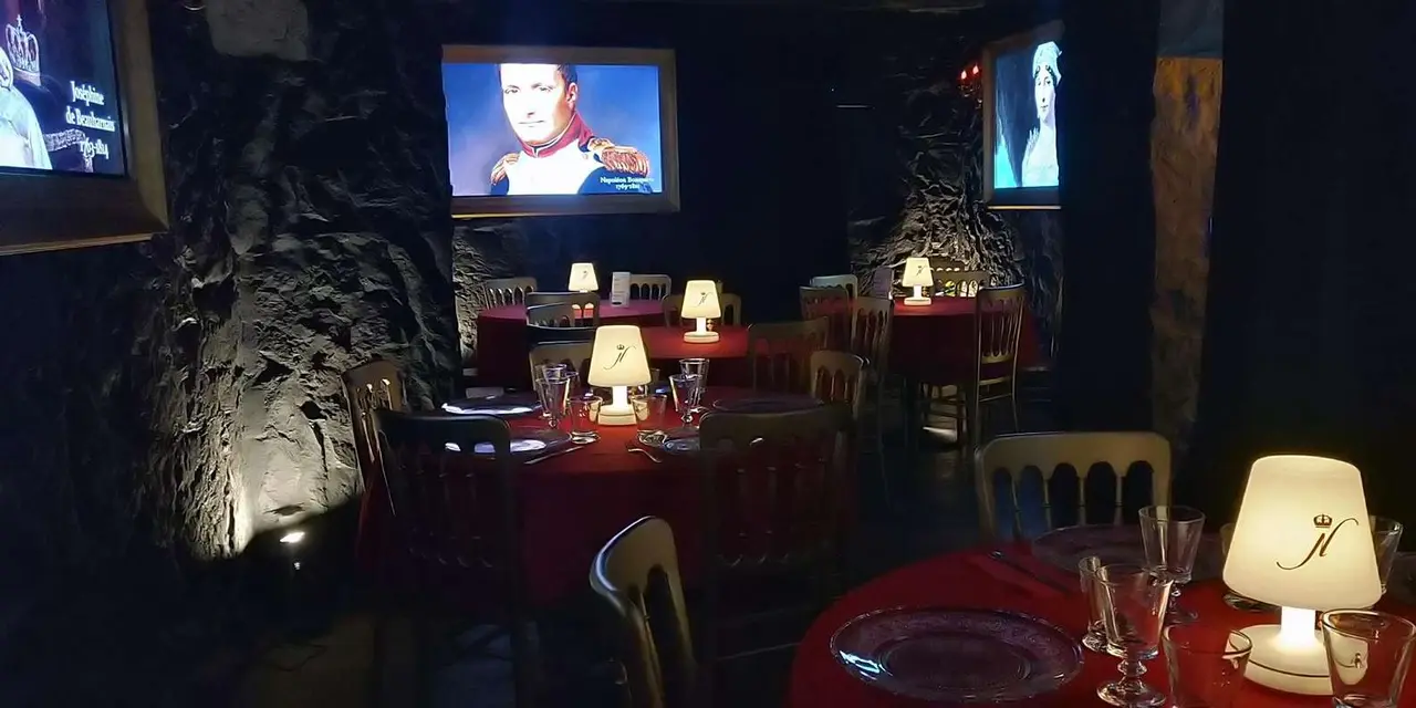 A dim vaulted room with dining tables and screens showing a portrait of Napoleon
