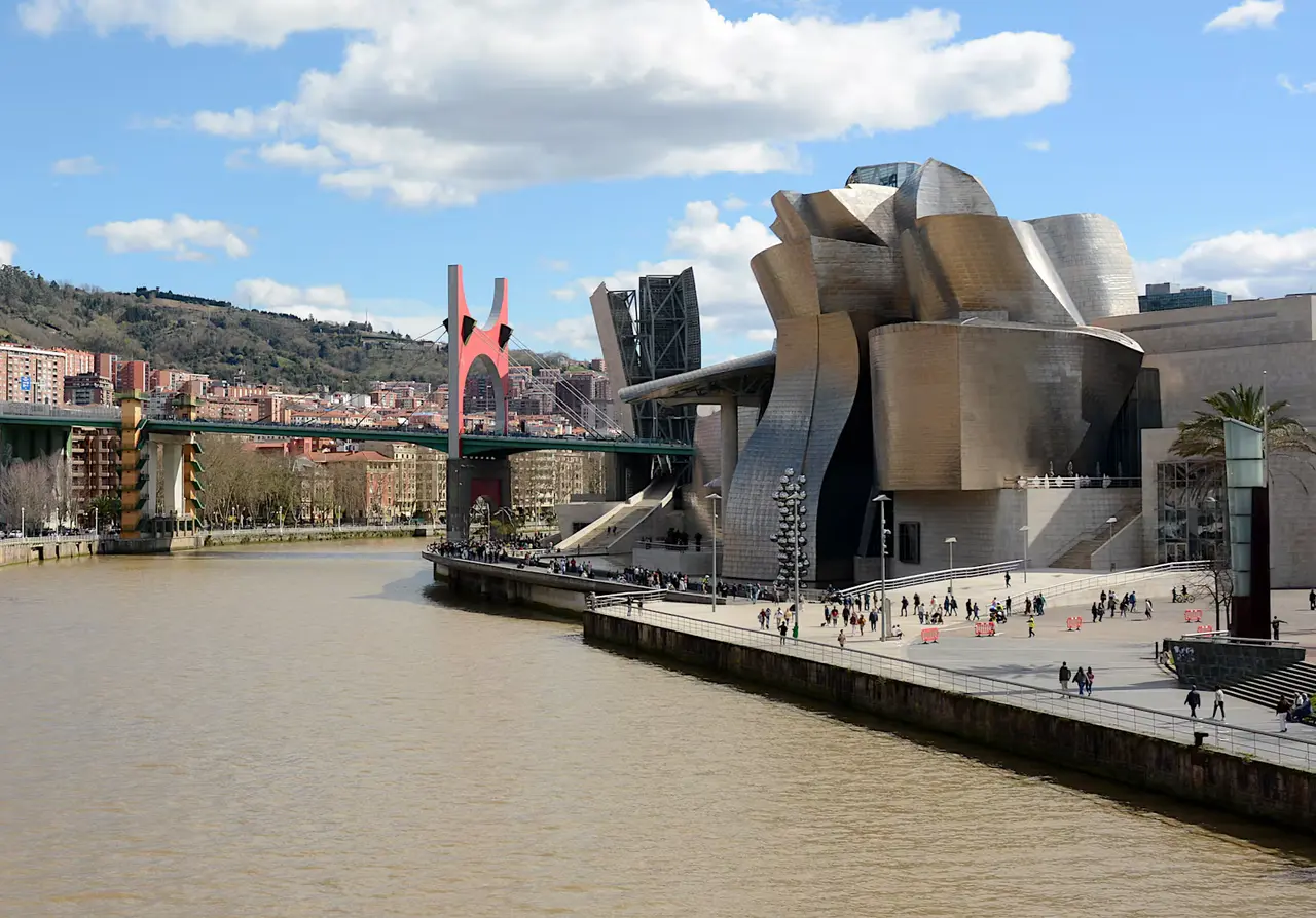The Guggenheim Museum Bilbao's curved metal walls beside the brown Nervión river, with the red arch of the La Salve bridge and a green hillside under a blue sky with white clouds