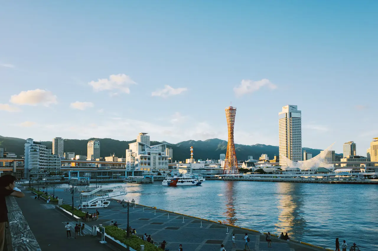 Kobe harbor on a clear evening: the red lattice Kobe Port Tower, the white sail-roofed Maritime Museum and Hotel Okura tower, with green mountains behind and people strolling the Harborland promenade