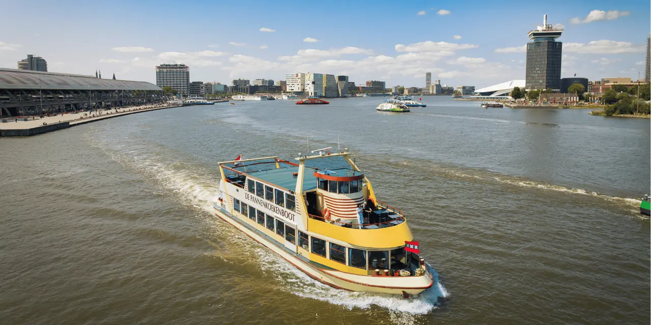 A yellow and cream pancake boat crossing the wide River IJ with Central Station's long roof on the left and the A'DAM Tower on the right