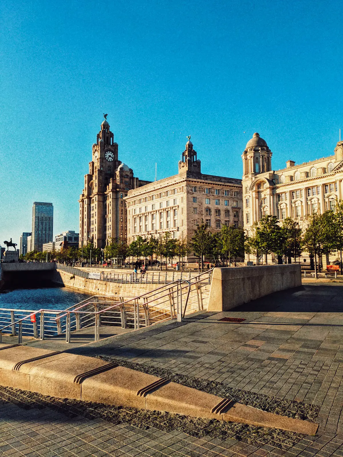 The Royal Liver Building, Cunard Building and Port of Liverpool Building on Liverpool's Pier Head under a clear blue sky, with the river-side promenade in front