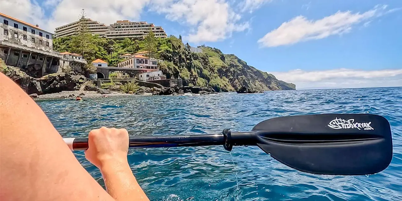 A hand gripping a kayak paddle above blue water, with a green cliff and buildings along the coast behind