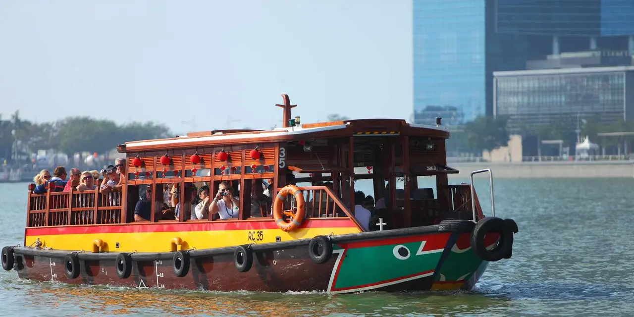 A red and green wooden bumboat carrying passengers on the Singapore River with a glass tower behind
