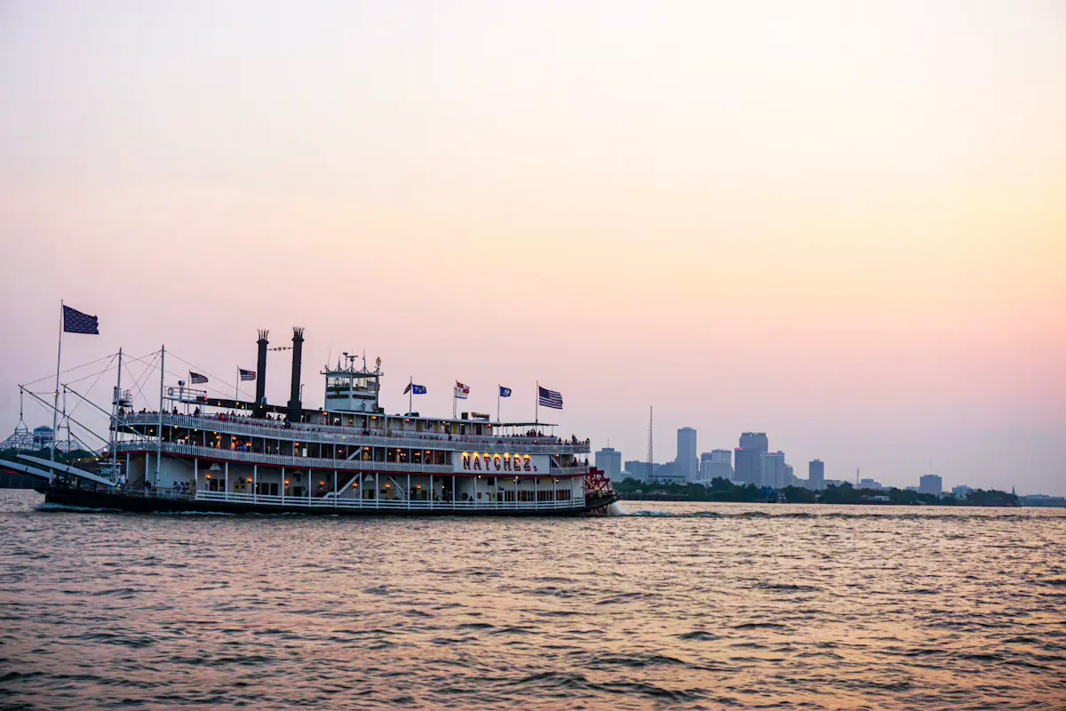 A paddlewheel steamboat with flags flying cruises the Mississippi at sunset, the downtown New Orleans skyline on the far bank under a pink sky