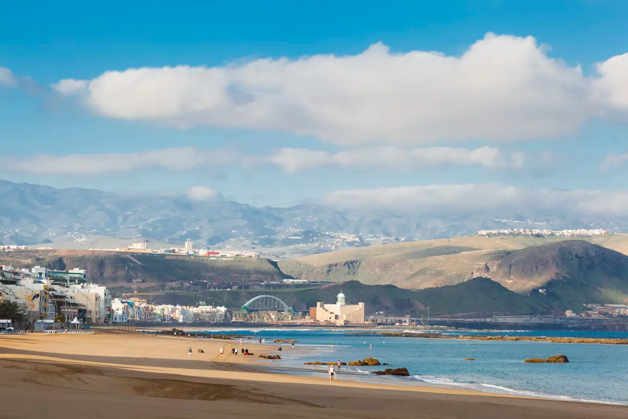 A wide sandy beach in Las Palmas de Gran Canaria with a few walkers at the water's edge, the Alfredo Kraus Auditorium on the shore and green mountains under a blue sky with white clouds