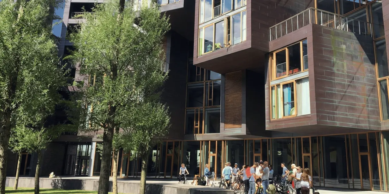 A tour group on bikes gathered at the foot of a modern apartment building of stacked copper-clad boxes with wooden-framed windows, trees in front
