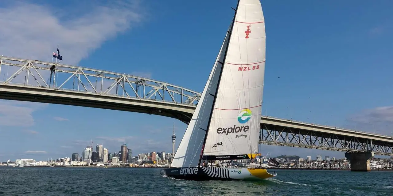 A racing yacht under full white sail passing beneath the Auckland Harbour Bridge, the city skyline behind