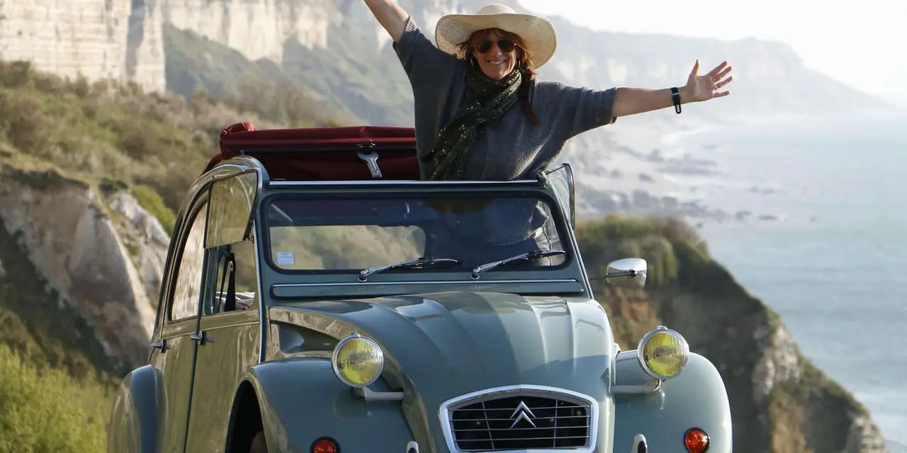 A woman with her arms raised standing through the roof of a grey-green Citroën 2CV on a cliff-top road above the sea