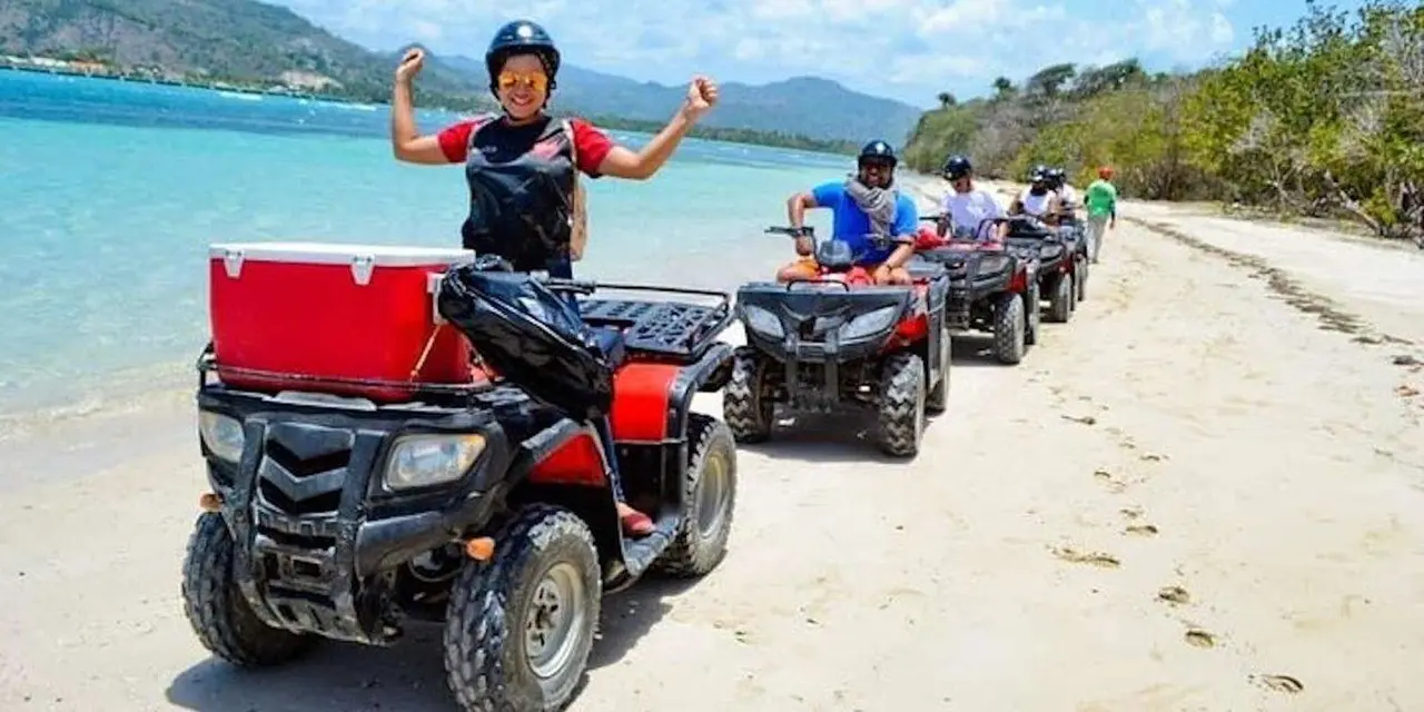 A rider raising both arms on a red ATV parked on a beach, with turquoise water and green hills behind