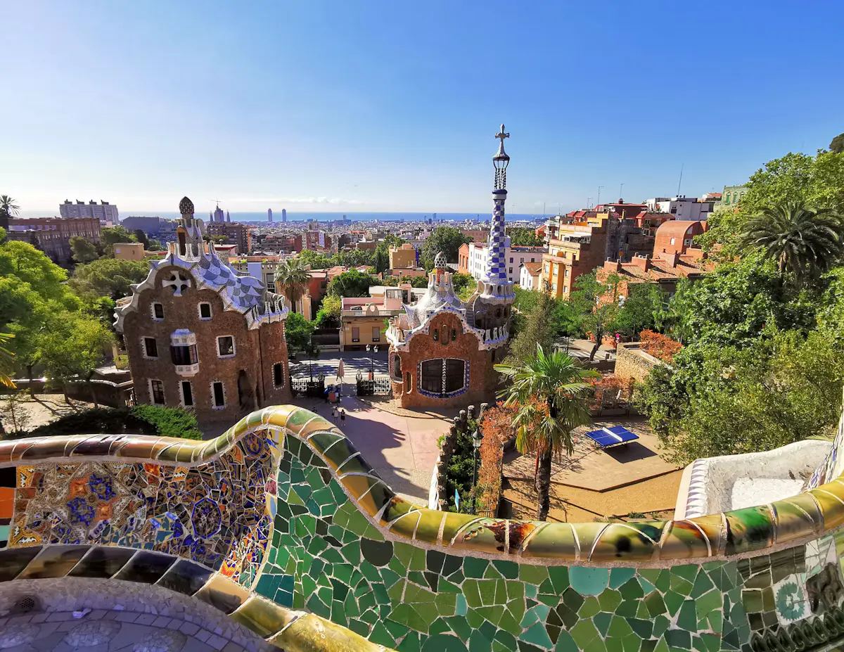 The colourful mosaic bench of Park Güell curling across the foreground, with the gingerbread-style gatehouses, rooftops of Barcelona and a strip of the Mediterranean under a clear blue sky