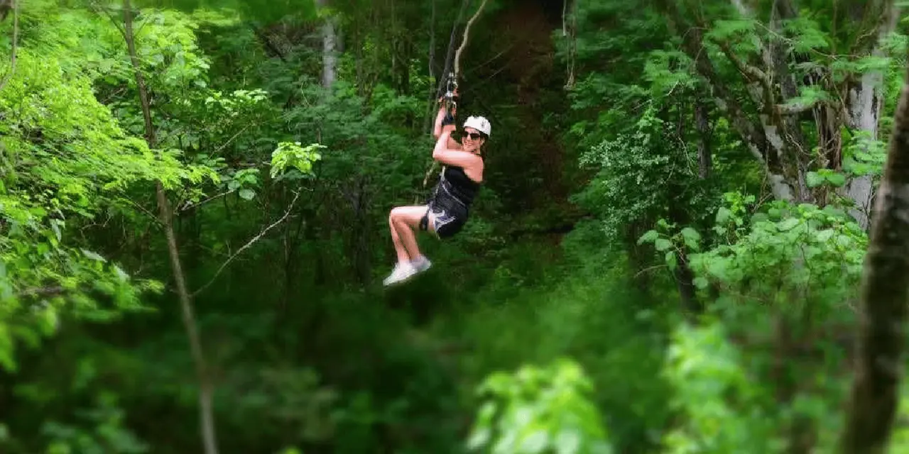 A woman in a helmet sliding along a zipline through green jungle foliage