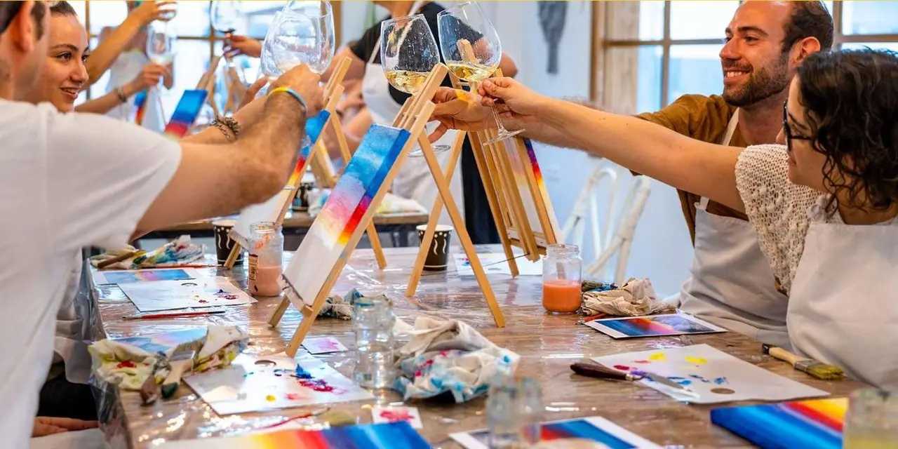 Hands raising wine glasses over easels and paint-covered tables