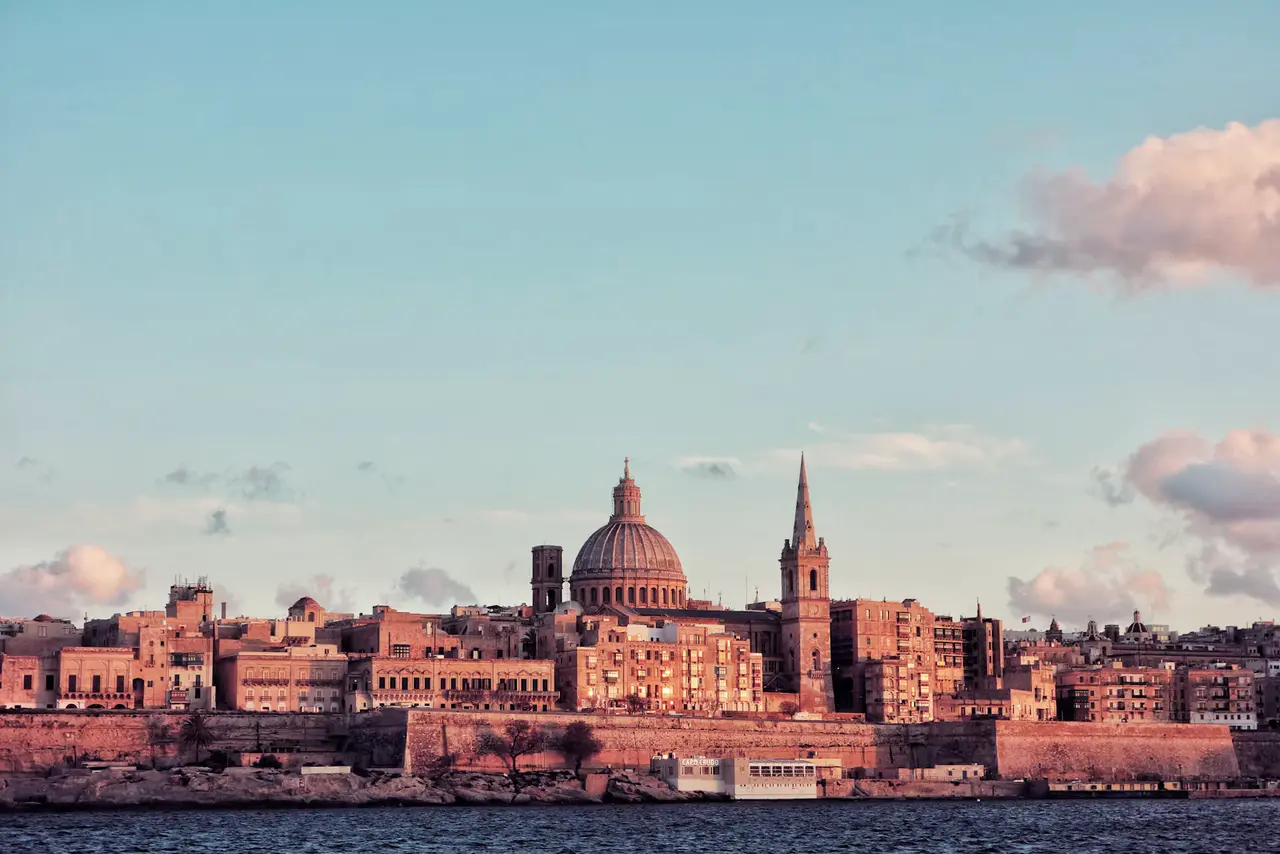 Valletta's skyline glowing pink at sunset across the harbour, a large dome and a tall church spire rising above the old bastion walls