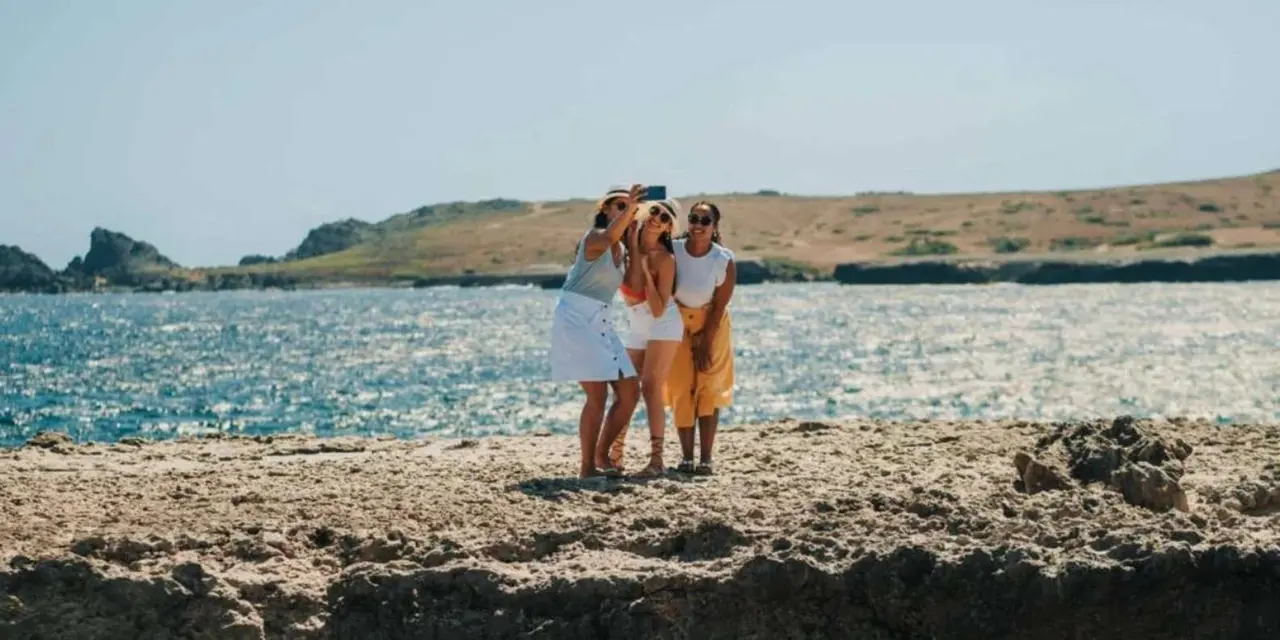 Three women taking a selfie together on a flat rocky shoreline with sparkling blue sea and brown hills behind