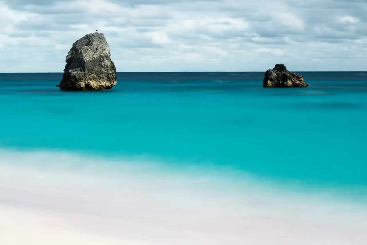 Two dark rock stacks rising from glassy turquoise water off a pale pink-white Bermuda beach under a soft grey sky